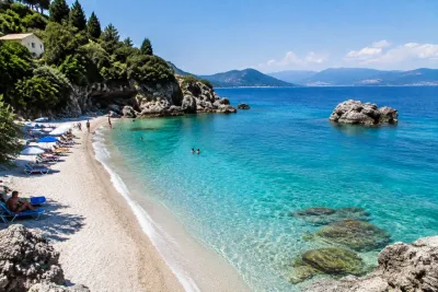 Turquoise water en kiezelstrand met ligstoelen, zwemmers en rotsige kustlijn in Corfu