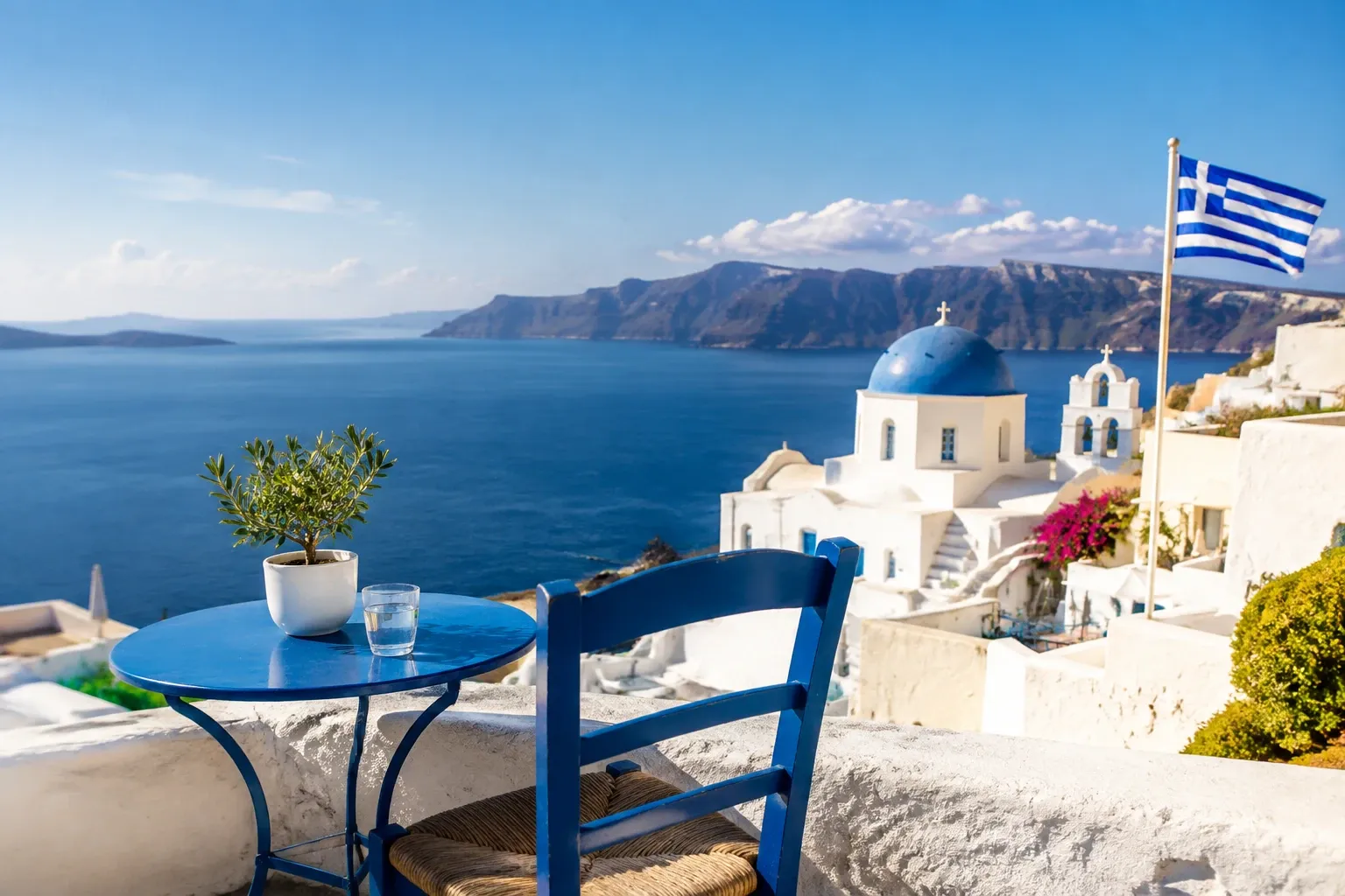 Terras met blauw tafeltje, stoel en olijfboompje, uitzicht op Santorini zee en kerk